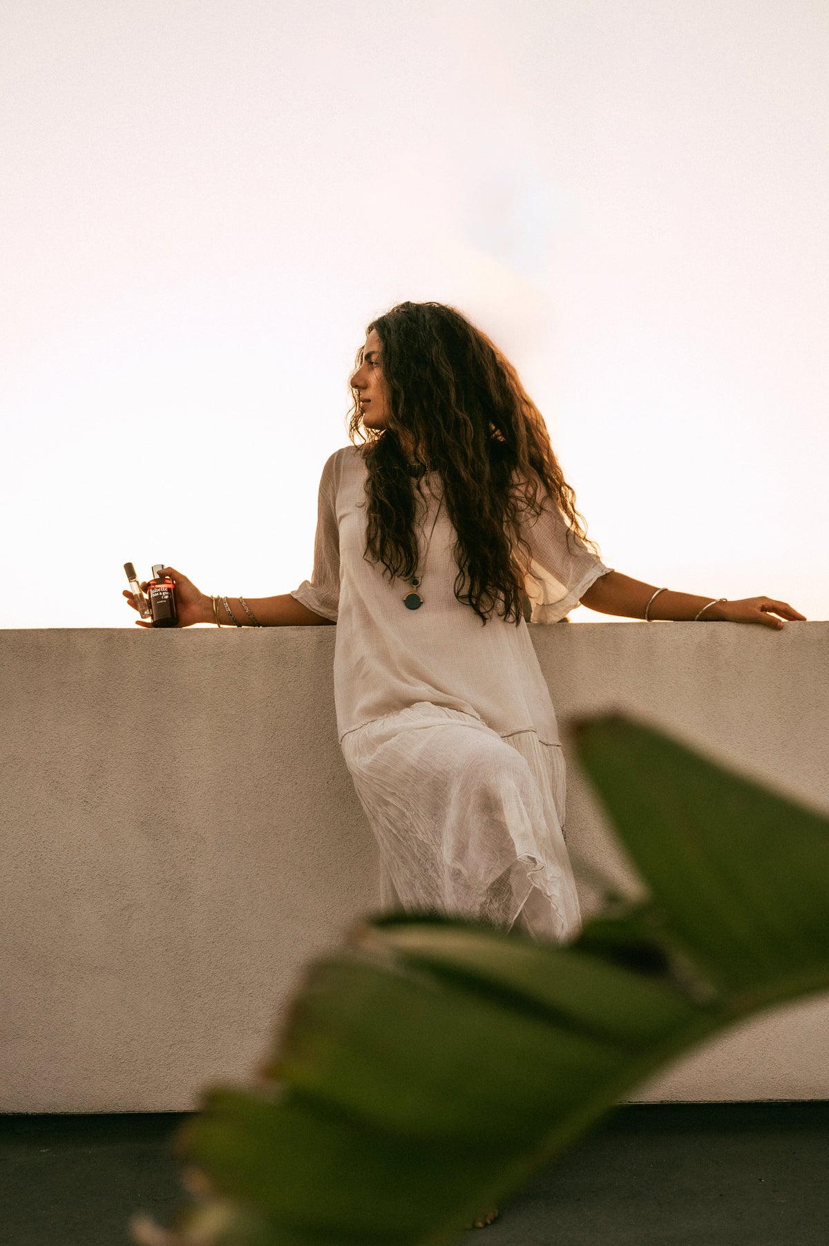 Model leaning on a rooftop wall at sunset holding a perfume oil roller